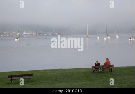 Couple resting on a bench overlooking the Gareloch in the mist, Scotland, with yachts in the background. Stock Photo