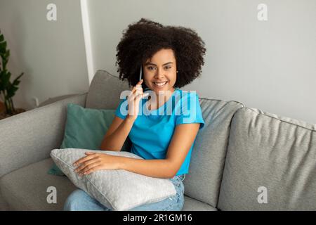 Good news, gossip. Glad pretty young black curly woman calling by smartphone, talking to friend on couch Stock Photo