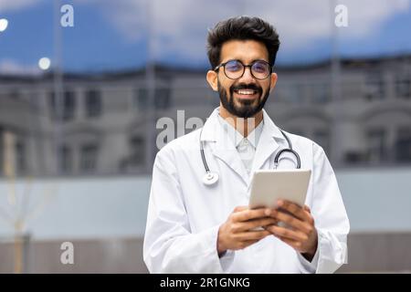 Portrait of happy and smiling hispanic doctor, man smiling and looking at camera, doctor using tablet computer for online consultation, outside of modern clinic. Stock Photo