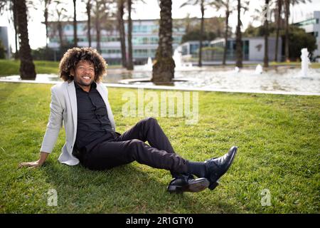 Happy black middle aged businessman sitting on grass in park, having break and feeling free, man escaping from work Stock Photo