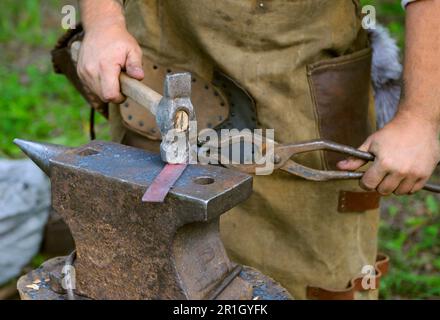 Blacksmiths hands holding forceps and hammer forging a metal billet, blade of a knife, on an anvil. Stock Photo