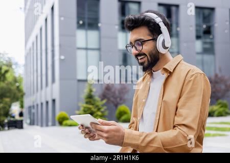 A young Inus man walks around the city in headphones, a businessman in a shirt watches an online video stream using a tablet computer, a programmer in a shirt outside an office building. Stock Photo