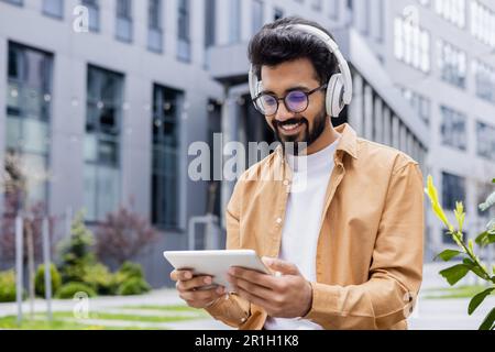 A young Inus man walks around the city in headphones, a businessman in a shirt watches an online video stream using a tablet computer, a programmer in a shirt outside an office building. Stock Photo