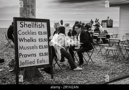 People eat traditional shellfish and seafood next to a price list board on the pebble beach at Brighton, East Sussex, UK. Brighton, a lively and vibra Stock Photo