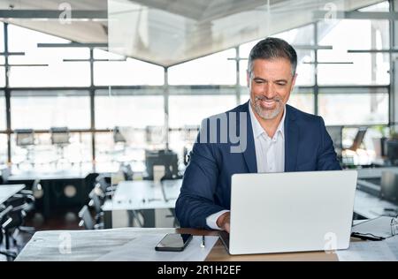 Smiling mature adult business man executive sitting at desk using laptop. Stock Photo