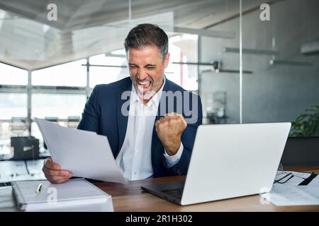 Happy middle aged business man holding documents celebrating success in office. Stock Photo