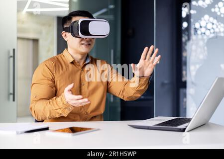 Successful businessman in online meeting uses vr glasses virtual reality, man communicates remotely with partners in metaverse, works inside office sitting at workplace with laptop. Stock Photo