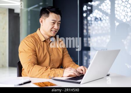 Cheerful and smiling Asian man working inside office, businessman in shirt and glasses using laptop, engineer developer programmer typing code programming software Stock Photo
