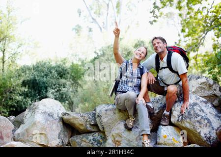 Taking a break while enjoying the view. A young woman pointing at something in the distance with her husband sitting next to her. Stock Photo
