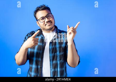 Indian young guy wearing eye glasses pointing finger aside on blue background Stock Photo