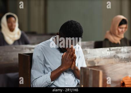 African American man praying with his eyes closed while sitting on bench in church Stock Photo