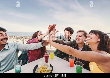 A diverse group of friends, including a non-binary person, celebrate their friendship with a high-five at an outdoor bar, sea and sky in the backgroun Stock Photo