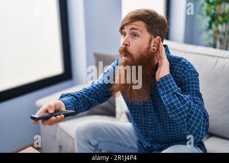 Young redhead man using deafness aid machine watching tv at home Stock Photo