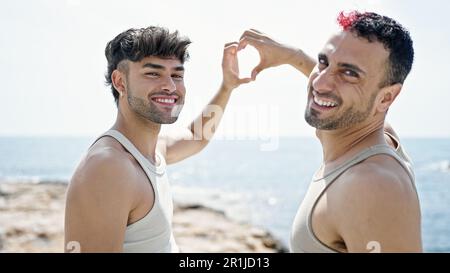 Two men couple hugging each other doing heart gesture at seaside Stock Photo