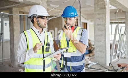 Two men builders talking on walkie talkie reading document at construction site Stock Photo