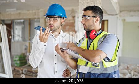 Two men builders and architect writing on document sending voice message by smartphone at construction site Stock Photo
