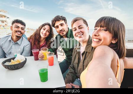 A multiracial group of friends takes a selfie at an outdoor bar table, with colorful drinks on the table, all smiling. Stock Photo