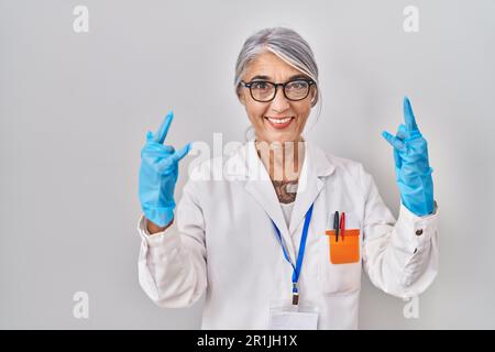 Middle age woman with grey hair wearing scientist robe shouting with crazy expression doing rock symbol with hands up. music star. heavy concept. Stock Photo