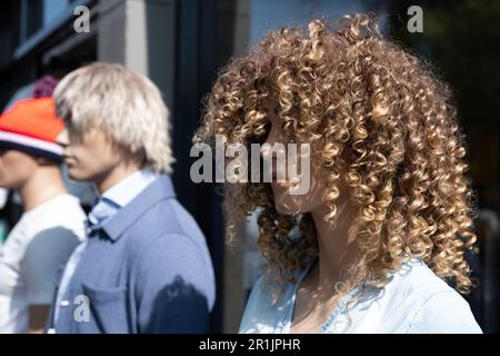Three plastic mannequins stand side by side in front of a fashion store facade. Focus on the right one wearing a curly wig Stock Photo