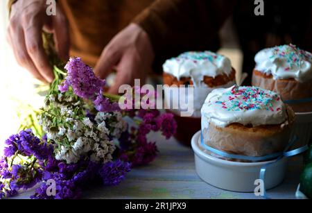 Three Easter cakes for the holiday are on the table. The hands of a man hold a bouquet of immortelle. Flowers in different colors Stock Photo