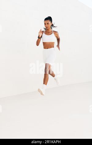 Full length of young slim woman sprinting outdoors at white wall. Fit woman running fast. Stock Photo