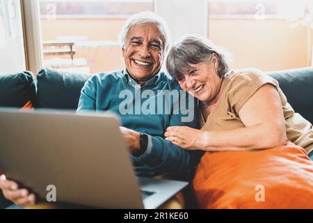 A joyful retired couple sits on the couch, with the man holding a laptop on his lap as they embrace, laughing heartily at online content. Stock Photo