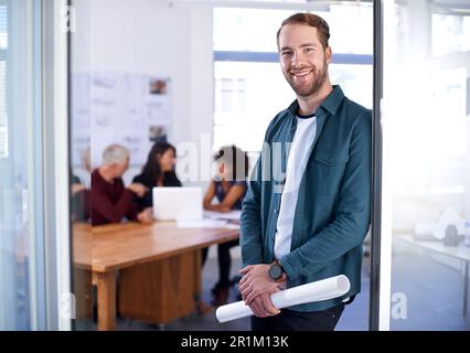 Theres nothing better than loving what you do. Portrait of a young architect holding blueprints with his colleagues working in the background. Stock Photo