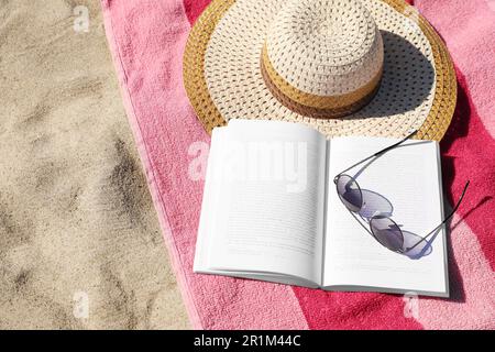 Open book, sunglasses, hat and striped towel on sandy beach, above view. Space for text Stock Photo