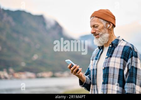 Older elder hipster man standing in nature park wearing earbud using phone. Stock Photo