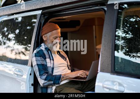 Happy older man traveler sitting in camper van using laptop in camping. Stock Photo