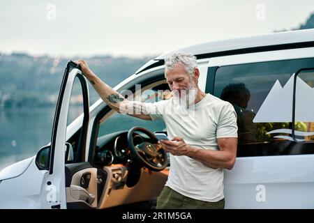 Mature man traveler standing near camper van using mobile in camping. Stock Photo