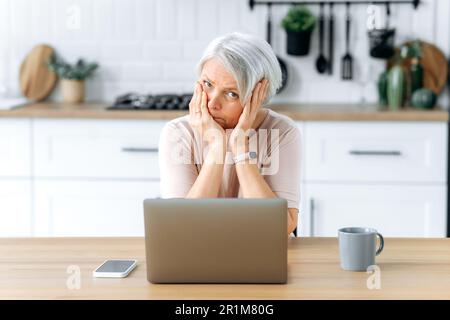 Puzzled exhausted mature caucasian gray haired woman, pensioner, sits at a table with laptop in kitchen, feels overworked and stressed, fatigue from working on a laptop, looks sadly at camera Stock Photo