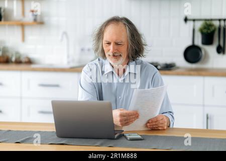 A positive mature gray haired caucasian man sits in the kitchen at a table with laptop, holds documents in his hands, does paperwork, reconciles data, analyzes the results of work Stock Photo