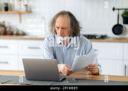Disappointed upset middle aged gray haired caucasian man sits in the kitchen at a table with laptop, holds documents in his hands, does paperwork, reconciles data, analyzes the results of work Stock Photo