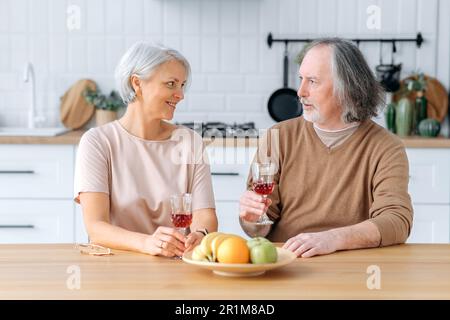 Middle aged caucasian spouses, sit at home in the kitchen, hold glasses of red wine in their hands, celebrate an important date, anniversary, look at glasses, smile joyfully. Happy pensioners Stock Photo
