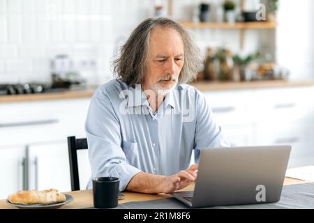 Focused senior caucasian gray haired man, sits at a desk with a laptop in kitchen at home, working online, checks and responds to email, browses news on the Internet, messaging Stock Photo