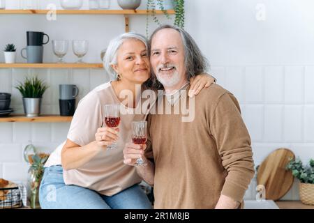 Photo of happy pensioners with wine. Middle aged caucasian spouses, sit at home in the kitchen, hold glasses of red wine in their hands, celebrate an important date, anniversary, smile at camera Stock Photo
