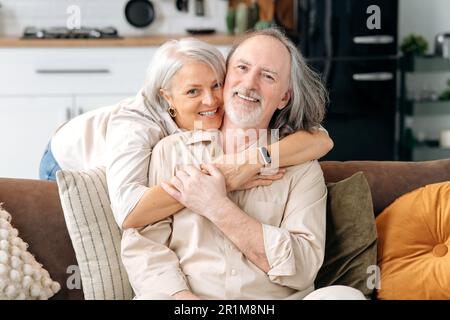 Positive happy caucasian mature married couple, gray haired spouses spend time together in a living room at home, woman embracing her husband on a sofa, they looking at camera, smiling Stock Photo