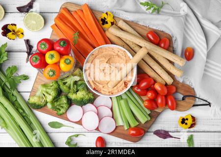 Board with delicious hummus, grissini sticks and fresh vegetables on white wooden background, flat lay Stock Photo