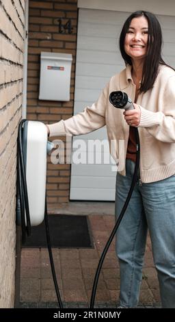 Young Central Asian woman plugging in a charging cable to charge her electric car. Sustainable and economic transportation concept. Stock Photo