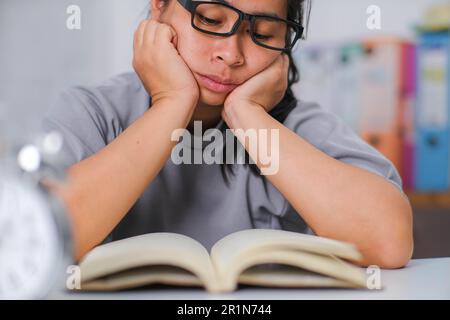 Tired Asian woman sitting at the desk reading a book in the living room. Stock Photo