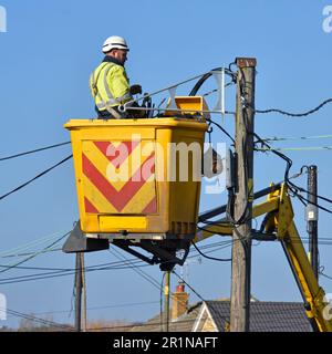 Electrician engineer in his workplace in a cherry picker hoist lift replacing all poles & overhead electricity cables in residential street England UK Stock Photo