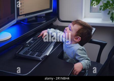 Happy baby is sitting at the computer keyboard next to the monitors. A smiling child with a desktop PC in a home living room. Kid aged about two years Stock Photo