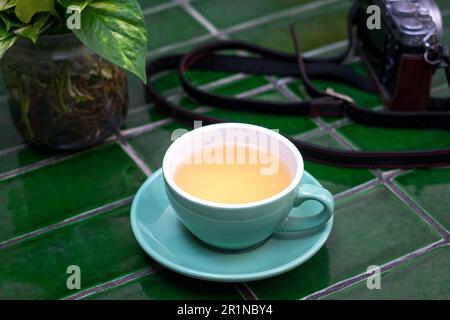Cup of chamomile tea with green plant and camera on top of table. Having a tea break concept. Stock Photo