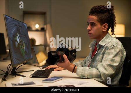 Young photographer sitting at his workplace with camera and retouching photo on computer Stock Photo