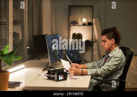 Young professional photographer working with photos in office while sitting at table with computer and camera Stock Photo