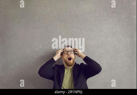 Portrait of happy shocked young man looking up with hands on head and mouth open Stock Photo