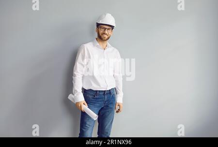 Happy smiling adult man architect wearing helmet and holding blueprint isolated on grey background. Stock Photo