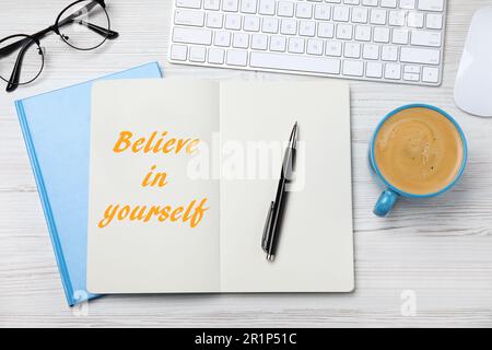 Phrase Believe in Yourself in notebook, computer keyboard and cup of coffee on white wooden desk, flat lay Stock Photo