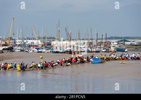 Runners taking part in the Maldon Mud Race in Maldon, Essex, UK, in the mud of the River Blackwater. Traditional charity event. Passing boats Stock Photo